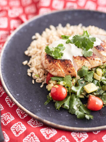 Sliced crispy chicken served over a bed of brown rice, accompanied by a zucchini and cherry tomato salad, topped with a dollop of sour cream and garnished with fresh coriander leaves.