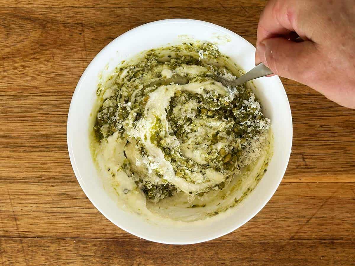 Greek yoghurt, pesto, Parmesan and lemon juice being stirred together in a bowl with a spoon.