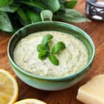 Fresh pesto dip in a green bowl with lemon slices and cheese on a wooden surface.