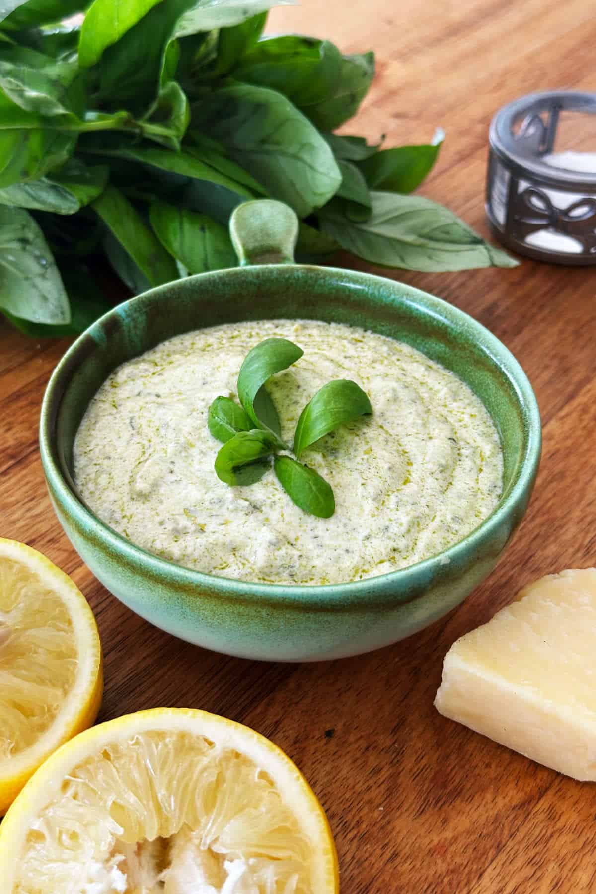 Fresh basil pesto dressing in a green bowl with lemon slices and cheese on a wooden surface.