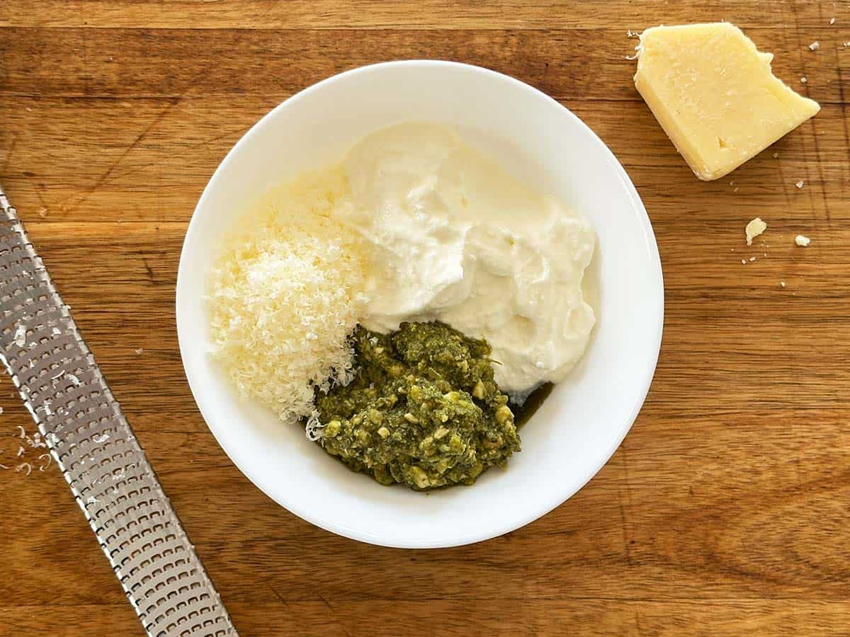 Step 1 in making basil pesto yogurt dressing - a bowl of greek yoghurt, parmesan cheese and basil pesto on a wooden board, ready to be mixed.