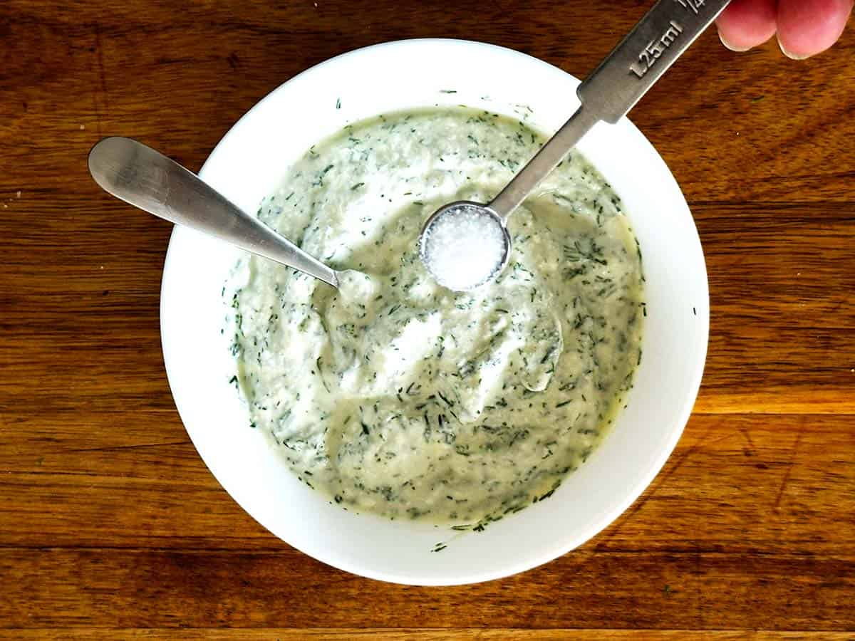 Small spoonful of salt held over a bowl of dill lemon yoghurt sauce for seasoning