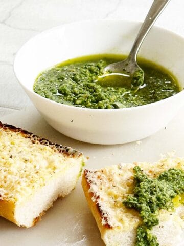 Basil pesto or pesto al basilico in a white bowl with two slices of garlic bread on a marble board, one slice topped with a dollop of pesto and a bite taken out of it.