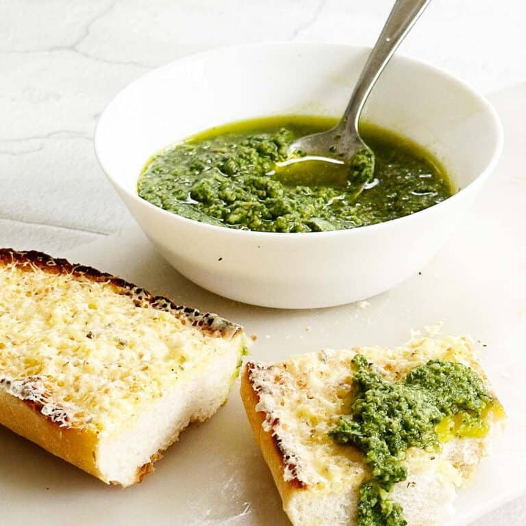 Basil pesto or pesto al basilico in a white bowl with two slices of garlic bread on a marble board, one slice topped with a dollop of pesto and a bite taken out of it.